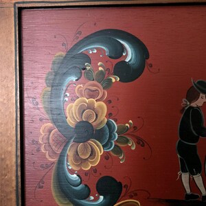 May include: Wooden framed folk art painting with a red background. The painting features a floral design in shades of yellow, blue, and green. A figure in black clothing is walking on the right side of the painting.