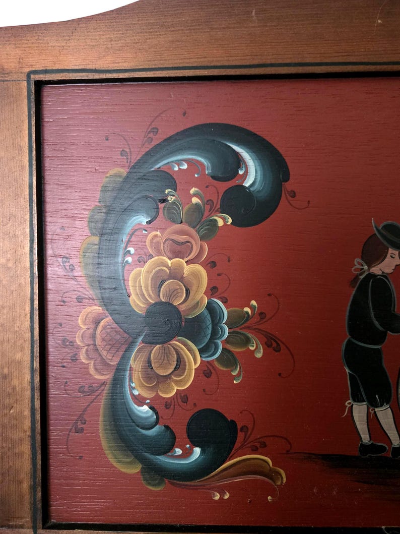 May include: Wooden framed folk art painting with a red background. The painting features a floral design in shades of yellow, blue, and green. A figure in black clothing is walking on the right side of the painting.
