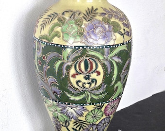 May include: A decorative ceramic vase with a cream-colored base and a flared, rounded shape. The vase features floral and foliage designs in green, purple, and gold. The central band has a dark green background with ornate patterns. The rim and base have gold accents.