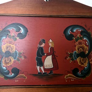 May include: Wooden wall art featuring a folk art painting of a couple in traditional attire holding hands. The red background is adorned with floral designs in blue, yellow, and brown. The frame is made of wood.