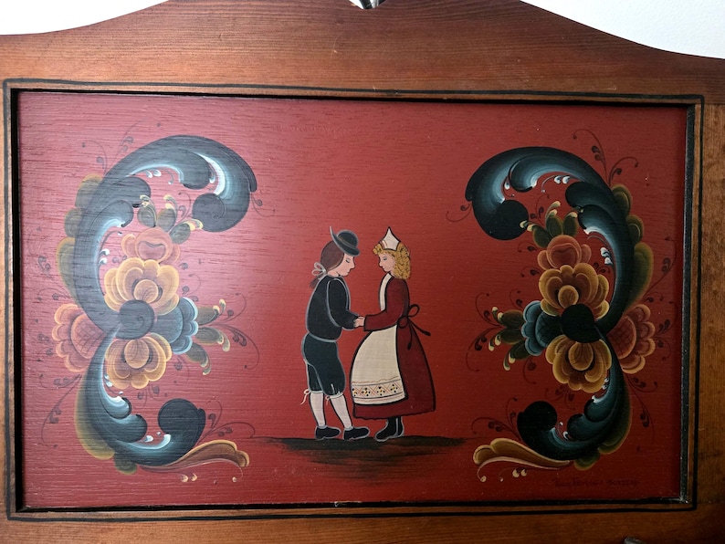 May include: Wooden wall art featuring a folk art painting of a couple in traditional attire holding hands. The red background is adorned with floral designs in blue, yellow, and brown. The frame is made of wood.