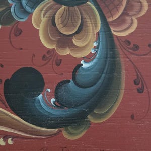 May include: Close-up of a wooden surface with a vibrant floral design in shades of yellow, blue, and black against a red background. The artwork features intricate details and a signature, adding a touch of artistry.