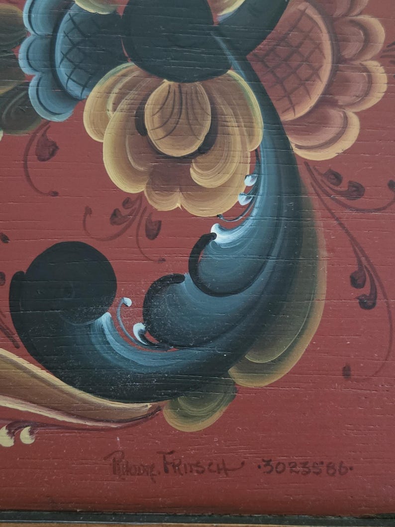 May include: Close-up of a wooden surface with a vibrant floral design in shades of yellow, blue, and black against a red background. The artwork features intricate details and a signature, adding a touch of artistry.
