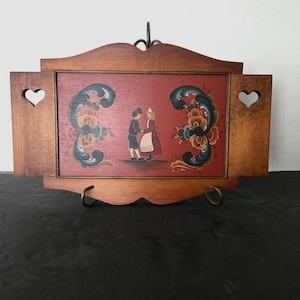 May include: Wooden wall art featuring a painted scene of two figures in traditional attire, set against a red background. The artwork is framed in brown wood with heart-shaped cutouts and decorative floral accents.