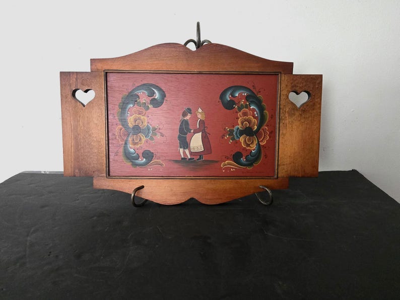May include: Wooden wall art featuring a painted scene of two figures in traditional attire, set against a red background. The artwork is framed in brown wood with heart-shaped cutouts and decorative floral accents.