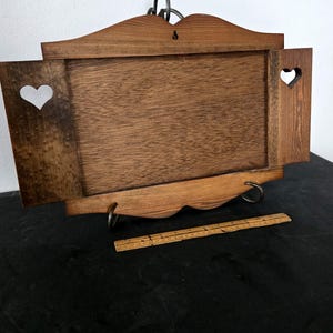 May include: Wooden wall-mounted tray with a rectangular center and two side panels, each featuring a heart-shaped cutout. The tray has a decorative top and bottom edge, with metal hanging hardware. A wooden ruler rests in front of the tray.