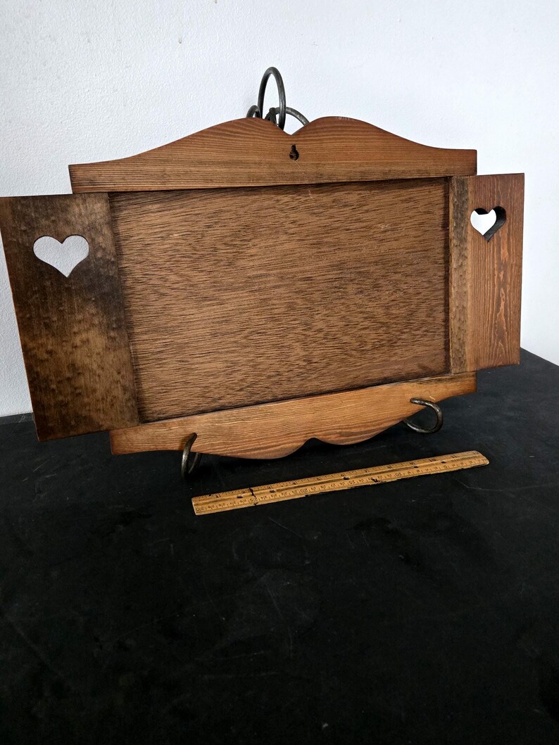 May include: Wooden wall-mounted tray with a rectangular center and two side panels, each featuring a heart-shaped cutout. The tray has a decorative top and bottom edge, with metal hanging hardware. A wooden ruler rests in front of the tray.