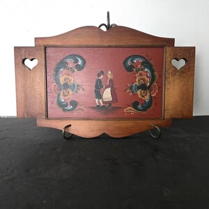 May include: Wooden framed artwork featuring a folk art painting of two figures in traditional attire. The painting is set against a red background and framed by a brown wooden frame with heart-shaped cutouts. The artwork is displayed on a black surface.