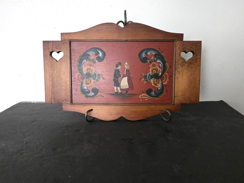 May include: Wooden framed artwork featuring a folk art painting of two figures in traditional attire. The painting is set against a red background and framed by a brown wooden frame with heart-shaped cutouts. The artwork is displayed on a black surface.