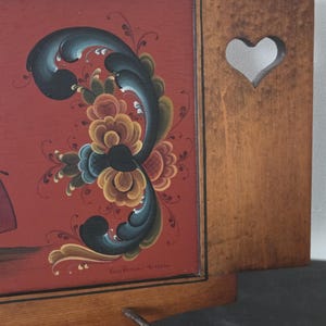 May include: Wooden wall decor with a red background and a floral design in shades of blue, yellow, and orange. The wooden frame has a heart-shaped cutout and a metal hook.