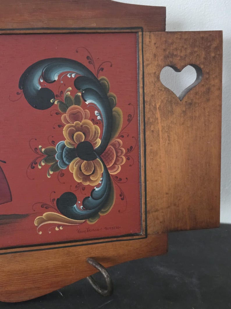 May include: Wooden wall decor with a red background and a floral design in shades of blue, yellow, and orange. The wooden frame has a heart-shaped cutout and a metal hook.