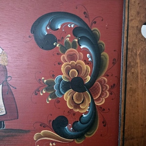 May include: A framed, folk art painting with a red background and a large, stylized floral design in shades of blue, yellow, and brown. The painting has a dark wooden frame. The artist's signature is visible in the lower right corner.