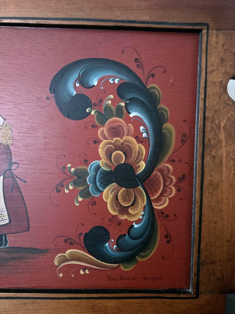 May include: A framed, folk art painting with a red background and a large, stylized floral design in shades of blue, yellow, and brown. The painting has a dark wooden frame. The artist's signature is visible in the lower right corner.