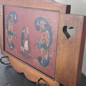 May include: Wooden serving tray with a painted folk art design. The tray features a red background with a couple in traditional clothing and floral accents. The tray has a heart-shaped cutout and metal handles.