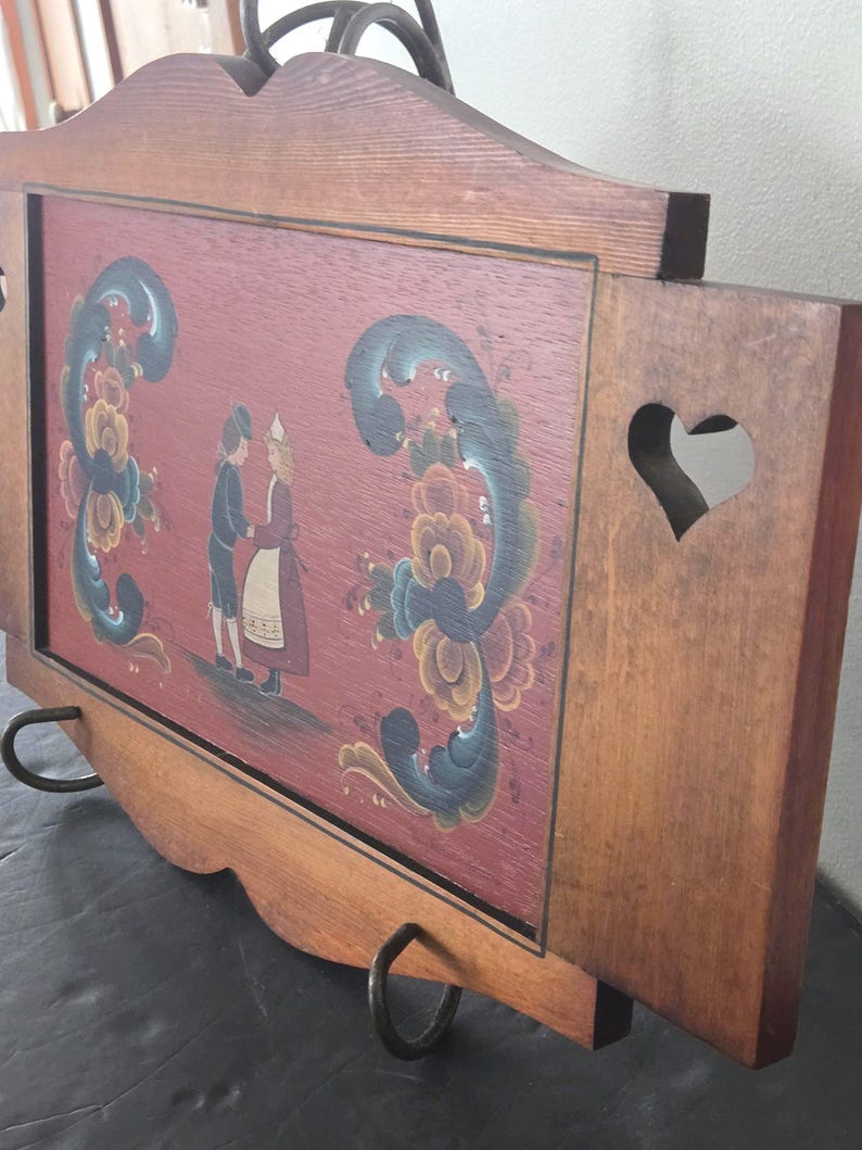 May include: Wooden serving tray with a painted folk art design. The tray features a red background with a couple in traditional clothing and floral accents. The tray has a heart-shaped cutout and metal handles.