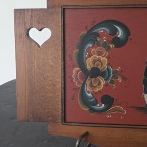 May include: Wooden wall-mounted decorative piece with a heart-shaped cutout and a painted floral design on a red background. The artwork features a dark floral pattern with yellow, blue, and green accents. A metal ring is attached to the bottom.