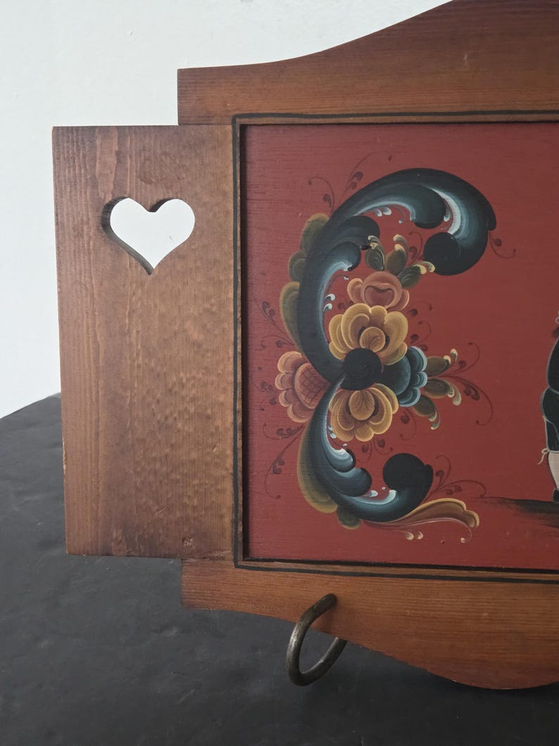 May include: Wooden wall-mounted decorative piece with a heart-shaped cutout and a painted floral design on a red background. The artwork features a dark floral pattern with yellow, blue, and green accents. A metal ring is attached to the bottom.