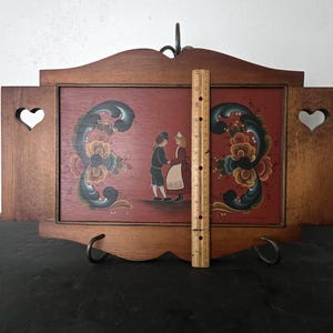 May include: Wooden wall art with a red background featuring a folk art painting of two figures dancing. The frame is brown with heart-shaped cutouts and decorative floral elements. A ruler shows the height is approximately 12 inches.