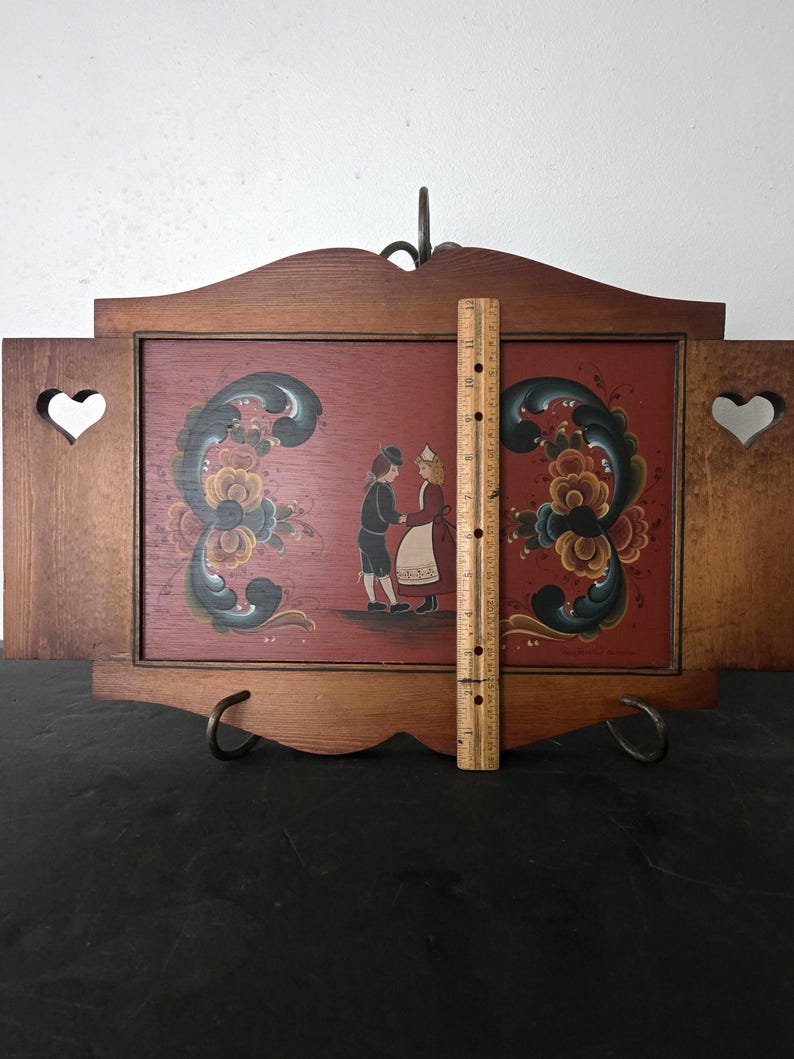 May include: Wooden wall art with a red background featuring a folk art painting of two figures dancing. The frame is brown with heart-shaped cutouts and decorative floral elements. A ruler shows the height is approximately 12 inches.