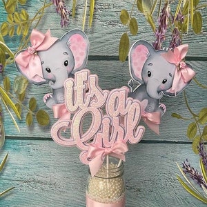 May include: Two pink and gray elephant figurines with pink bows and glitter accents are arranged in a glass jar with white beads and a pink ribbon. The text "it's a girl" is displayed in pink glitter letters.