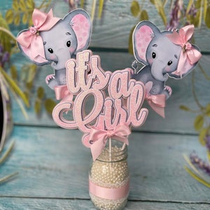 May include: It's a Girl centerpiece with two gray elephant figurines wearing pink bows and a pink glitter 'It's a Girl' sign. The centerpiece is in a clear glass jar with a pink ribbon and white beads.