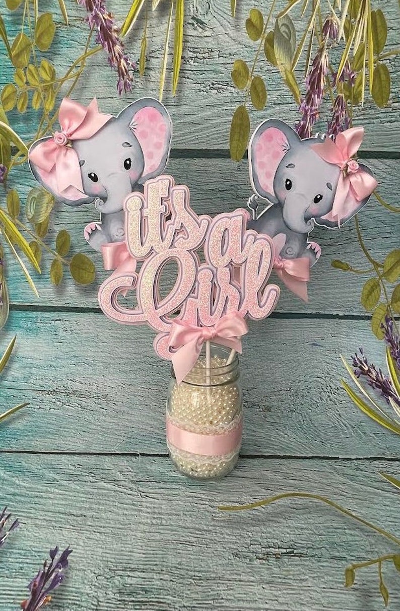 May include: Two pink and gray elephant figurines with pink bows and glitter accents are arranged in a glass jar with white beads and a pink ribbon. The text "it's a girl" is displayed in pink glitter letters.