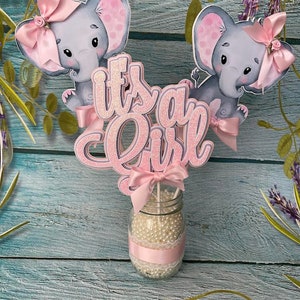 May include: It's a Girl centerpiece with two pink elephant figurines, a pink bow, and glitter lettering. The centerpiece is in a clear glass jar filled with white beads and a pink ribbon.