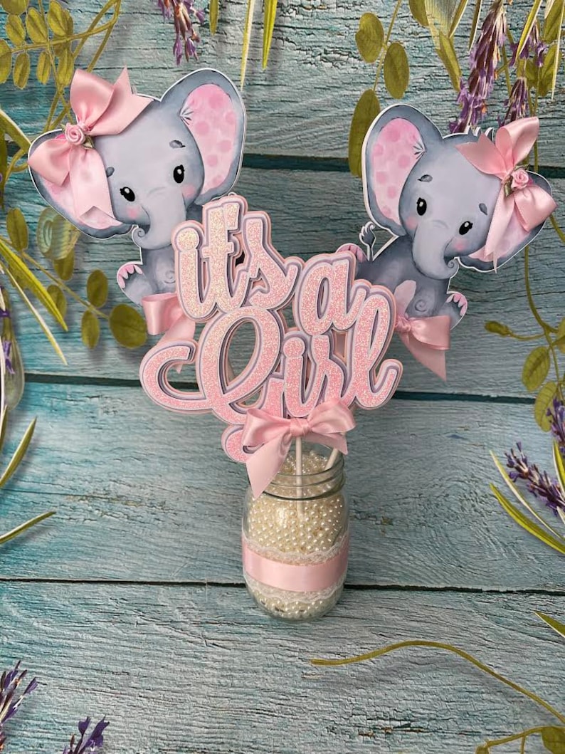 May include: It's a Girl centerpiece with two pink elephant figurines, a pink bow, and glitter lettering. The centerpiece is in a clear glass jar filled with white beads and a pink ribbon.