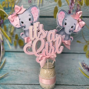 May include: Pink and gray elephant baby shower centerpiece with the words "It's a Girl" in pink glitter letters.