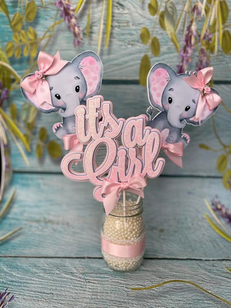May include: Pink and gray elephant baby shower centerpiece with the words "It's a Girl" in pink glitter letters.
