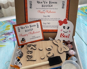 May include: A craft kit for painting a ghost with the words "You've Been Booed!" and "Happy Halloween". The kit includes a wooden ghost cutout, paint, a paintbrush, and a sponge brush. The kit is packaged in a white box with orange and black tissue paper.