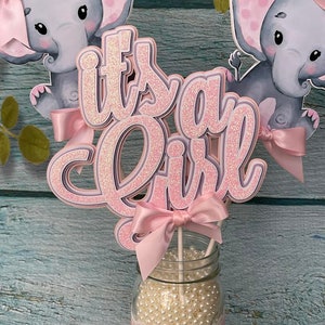 May include: Pink and gray elephant themed centerpiece with the words "it's a girl" in pink glitter letters. The centerpiece is in a clear glass jar with white beads and a pink ribbon.