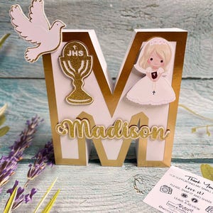 May include: White wooden letter M with gold trim, decorated with a white dove, a gold chalice with the letters JHS, and a girl in a white dress. The letter is decorated with the name Madison in gold glitter letters.