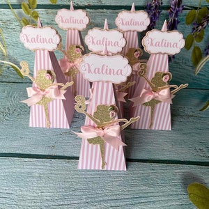 May include: Six pink and white striped cone-shaped party favors with a ballerina silhouette and the name "Kalina" on each. The cones are decorated with gold glitter and pink bows.