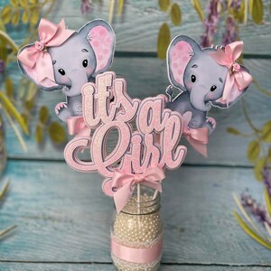 May include: Pink and gray elephant baby shower centerpiece with the words "It's a Girl" in pink glitter letters.