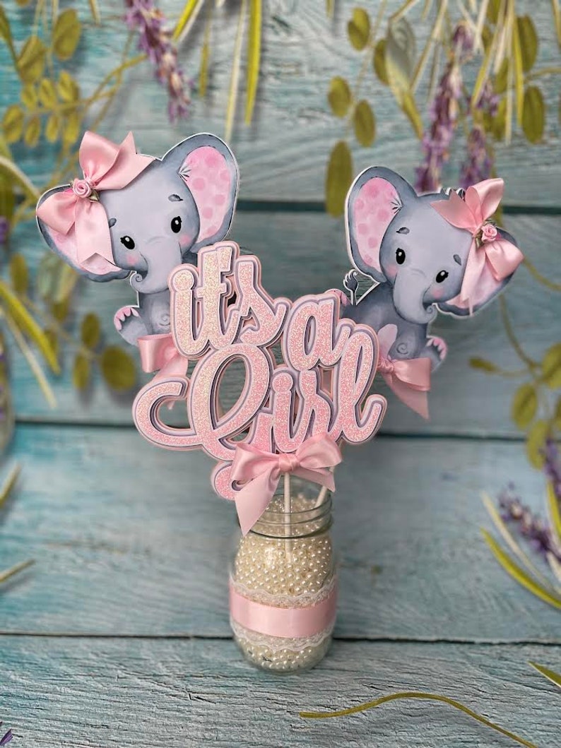 May include: Pink and gray elephant baby shower centerpiece with the words "It's a Girl" in pink glitter letters.