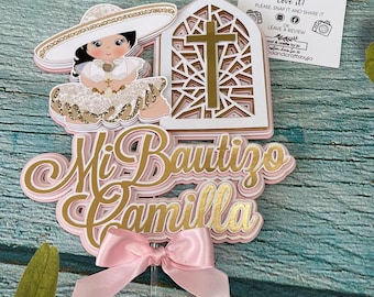 May include: A pink and gold cake topper with the words "Mi Bautizo Camilla" and a small girl wearing a white dress and a sombrero. The topper is decorated with a pink bow and a church window with a cross.