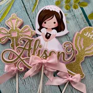 May include: Gold and pink cake toppers with a cross, the name Alison, a chalice with the letters JHS, and a girl in a white dress holding a bible.