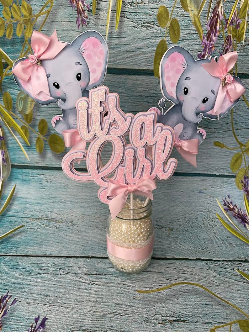 May include: It's a Girl centerpiece with two pink elephant figurines, a pink bow, and glitter lettering. The centerpiece is in a clear glass jar filled with white beads and a pink ribbon.