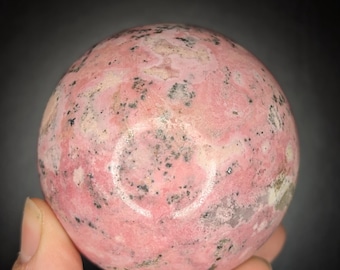 May include: A pink and gray sphere, held in a hand, against a dark background. The sphere has a mottled appearance with varying shades of pink and gray, and small black speckles. The surface appears smooth and polished.