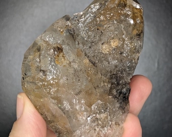 May include: A raw, natural quartz crystal with a smoky, translucent appearance. The crystal has a rough, textured surface with areas of clear and brown coloration. The overall shape is irregular, with sharp edges and a pointed tip. The crystal is held in a hand.