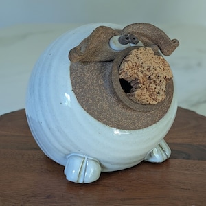May include: A whimsical ceramic pig-shaped container. The pig is primarily white with brown accents on the snout, ears, and around the opening. A cork stopper is visible in the mouth. The container has small, rounded feet.