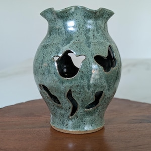 May include: A handcrafted, teal-colored ceramic vase with a ruffled rim and cut-out designs. The vase has a speckled finish and a dark interior, creating a contrast with the exterior. The vase is displayed on a wooden surface.