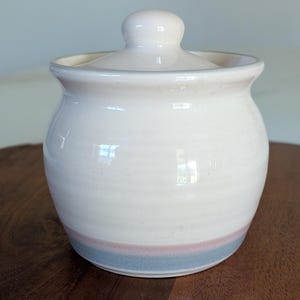 May include: A small, off-white ceramic jar with a matching lid. The jar has a rounded shape with a subtle pink and blue stripe near the base. The lid has a small, rounded knob.