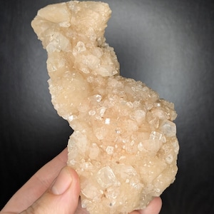 May include: A cluster of light beige crystals with a rough, textured surface. The crystals are held in a hand against a dark background. The mineral formation has a natural, organic shape, with many small, sparkling crystal points.