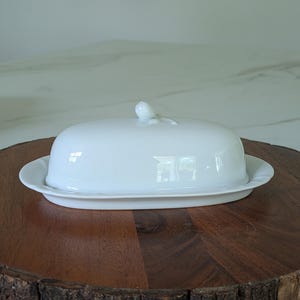 May include: A white ceramic butter dish with a lid, set on a wooden serving board. The dish has an oval shape with a small handle on the lid. The wooden board has a natural, rustic edge.