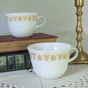 May include: Two white ceramic teacups with a gold floral pattern around the rim. The cups have a rounded shape with a handle. The cups are sitting on a wooden surface next to a gold candlestick.