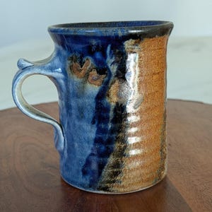 May include: A handmade ceramic mug with a unique design. The mug features a blue and brown glaze, with a textured surface. The handle is curved and attached to the side of the mug. The mug is approximately 4 inches tall.