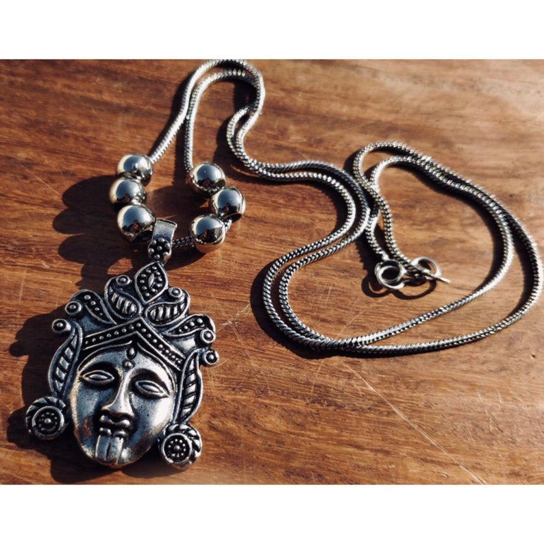 May include: A silver necklace with a pendant featuring a detailed face with a crown. The necklace has a chain with round beads.