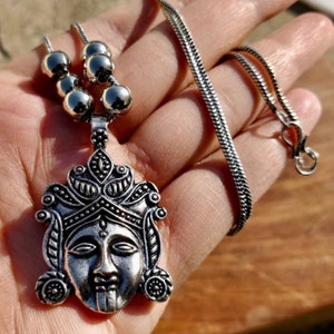 May include: Silver necklace with a detailed pendant featuring a face with a crown. The pendant is attached to a silver chain with round beads.